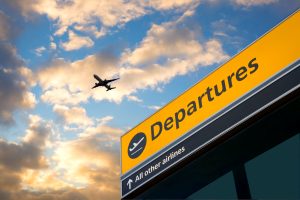 A stock photo of a departures sign at Heathrow Airport with a silhouetted plane in the sky behind it