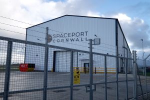 A photo of a hanger or warehouse with a sign that says 'Spaceport Cornwall' taken through a security fence